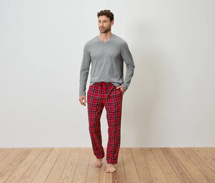 Un homme portant un t-shirt gris à manches longues et un pantalon de pyjama à carreaux rouges se tient sur un plancher en bois.