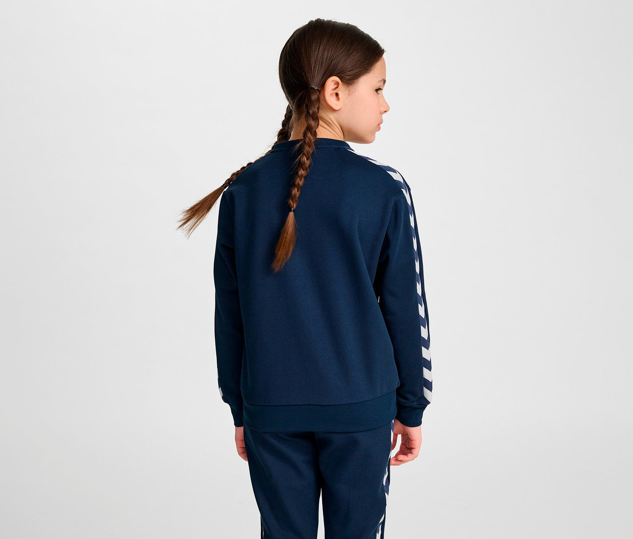 Une fille avec des tresses, vue de dos, porte une veste à fermeture éclair HUMMEL HMLJR Archive Reg Poly, bleue.