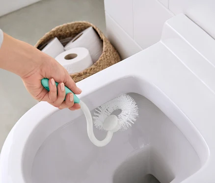 Une femme nettoie les toilettes avec une brosse blanche à manche turquoise. Un panier de papier toilette est visible à l'arrière-plan.