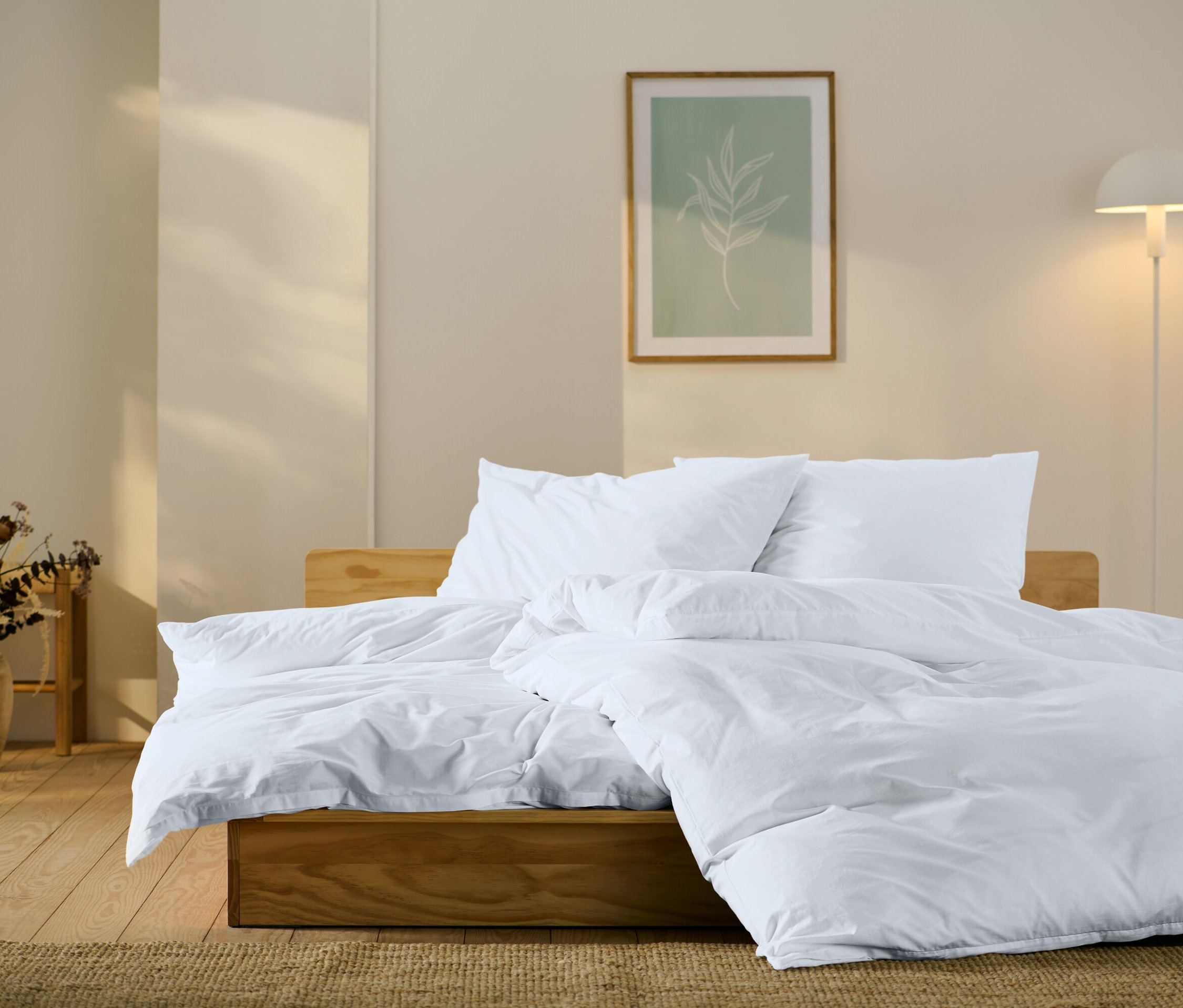 Lit en bois avec des draps, des oreillers et une couette blancs. Au mur, un tableau avec un motif de feuille et une lampe.