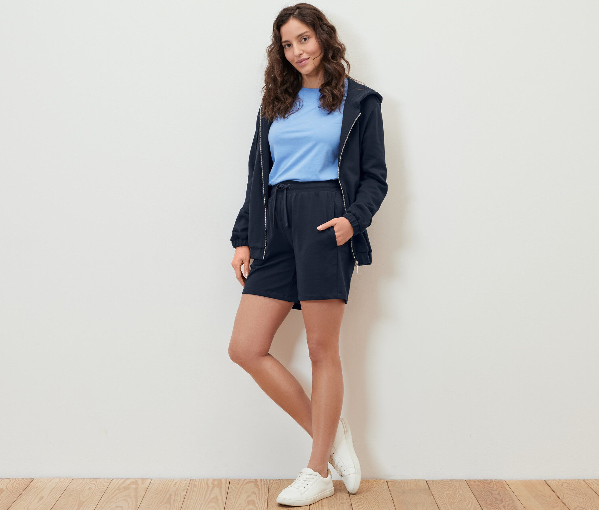 Femme posant en sweatshort bermuda, t-shirt bleu et sweat à capuche zippé.