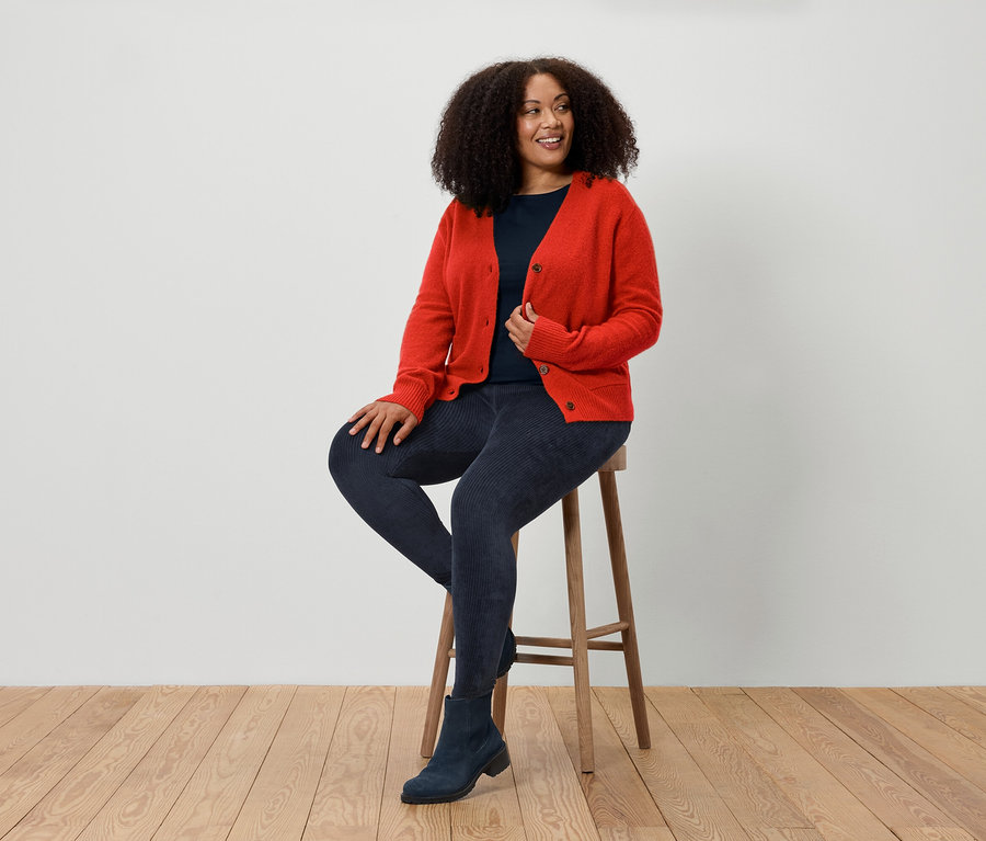 Une femme assise sur un tabouret porte un cardigan rouge, un jean bleu et des bottes bleues.