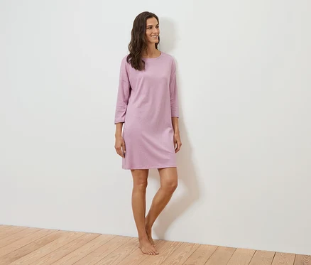 Une femme se tient debout et sourit, elle porte une robe violette à manches trois-quarts.