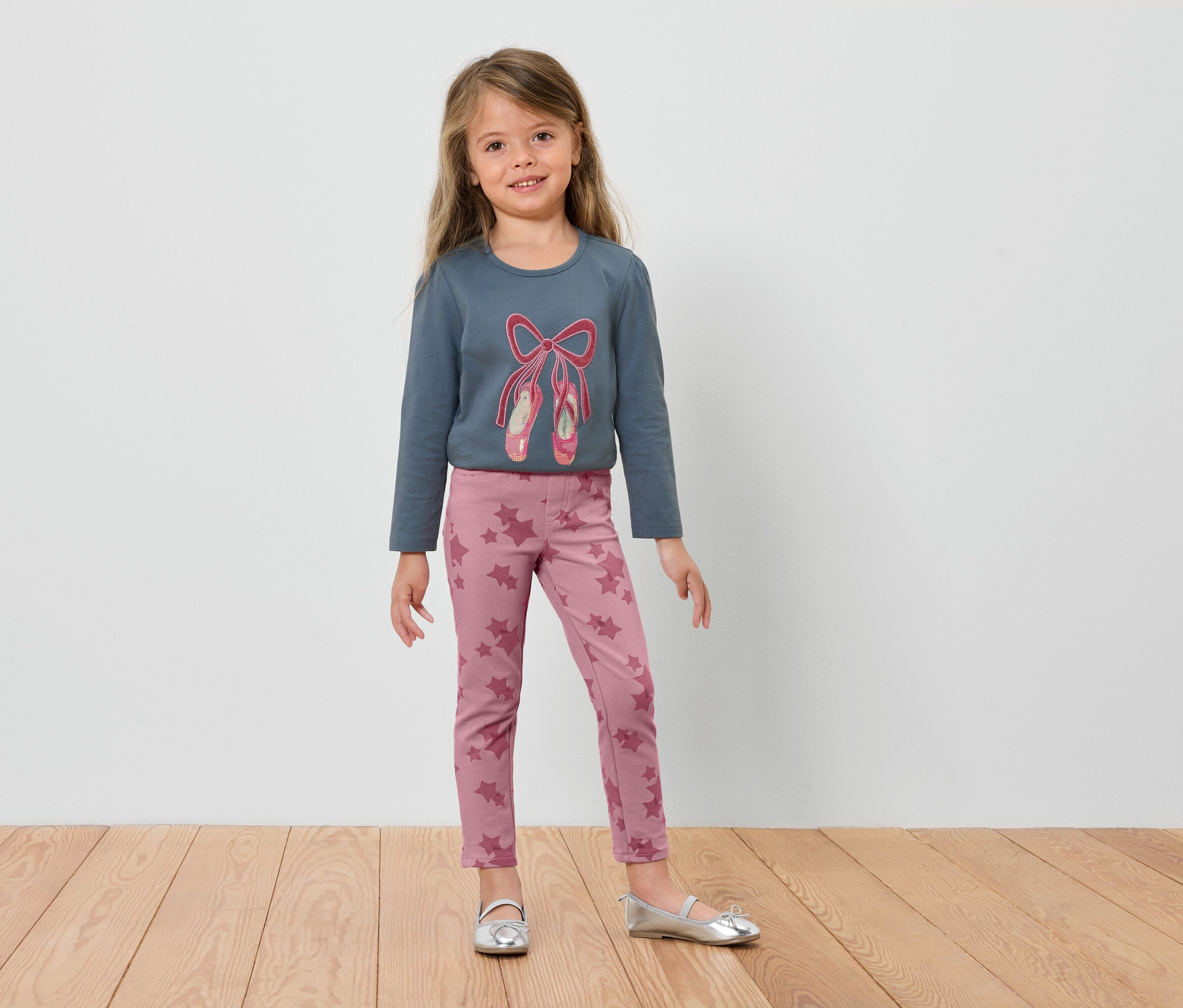 Une fillette portant un t-shirt bleu à manches longues avec des chaussons de ballet et un legging rose, se tenant sur un plancher en bois.