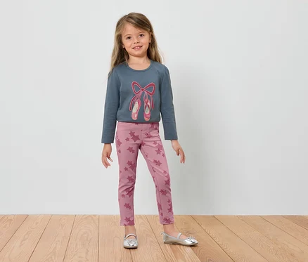 Une fillette portant un t-shirt bleu à manches longues avec des chaussons de ballet et un legging rose, se tenant sur un plancher en bois.