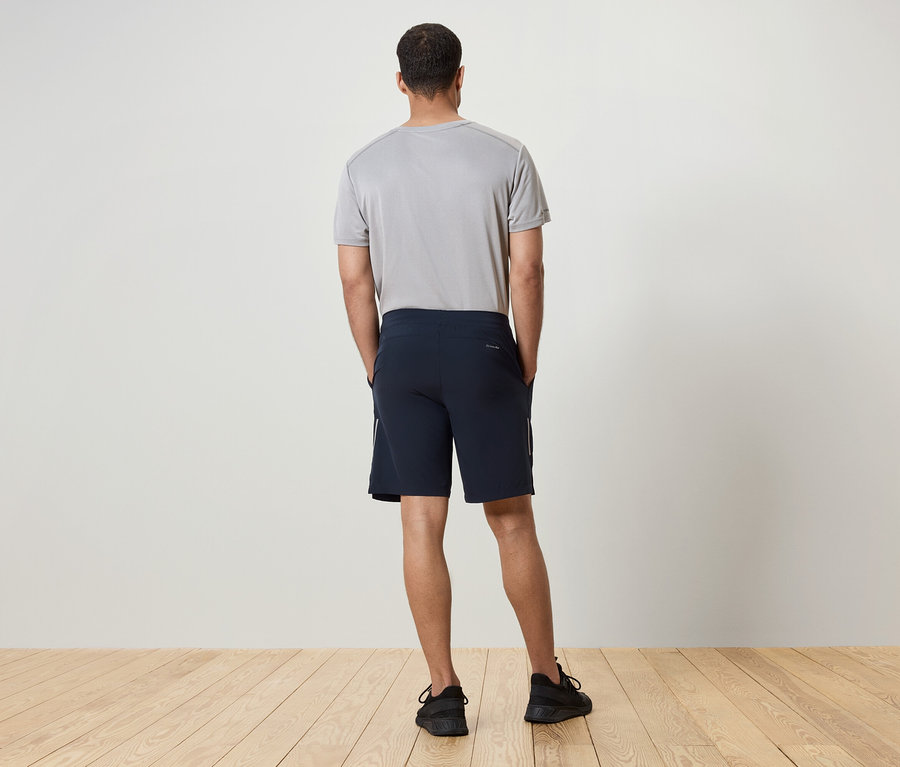 Un homme dos à la caméra porte un t-shirt gris, un short bleu marine et des baskets noires.