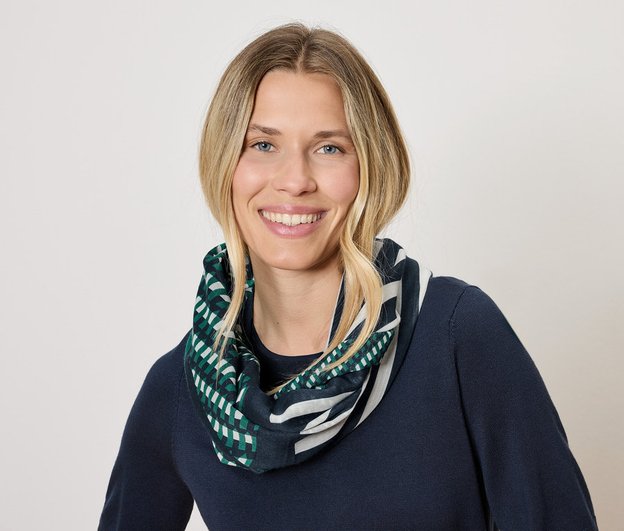 Une femme aux cheveux blonds et souriante porte un pull bleu marine et un foulard à motifs géométriques verts et blancs.