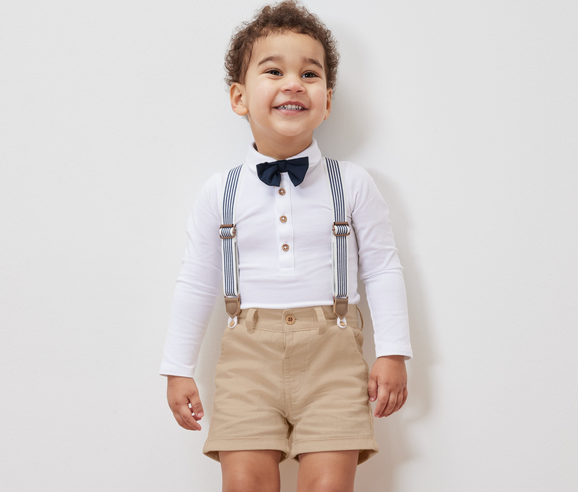 Petit garçon souriant portant une chemise blanche à manches longues, un nœud papillon, des bretelles et un short pour enfant.
