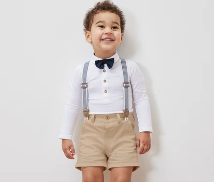 Petit garçon souriant portant une chemise blanche à manches longues, un nœud papillon, des bretelles et un short pour enfant.