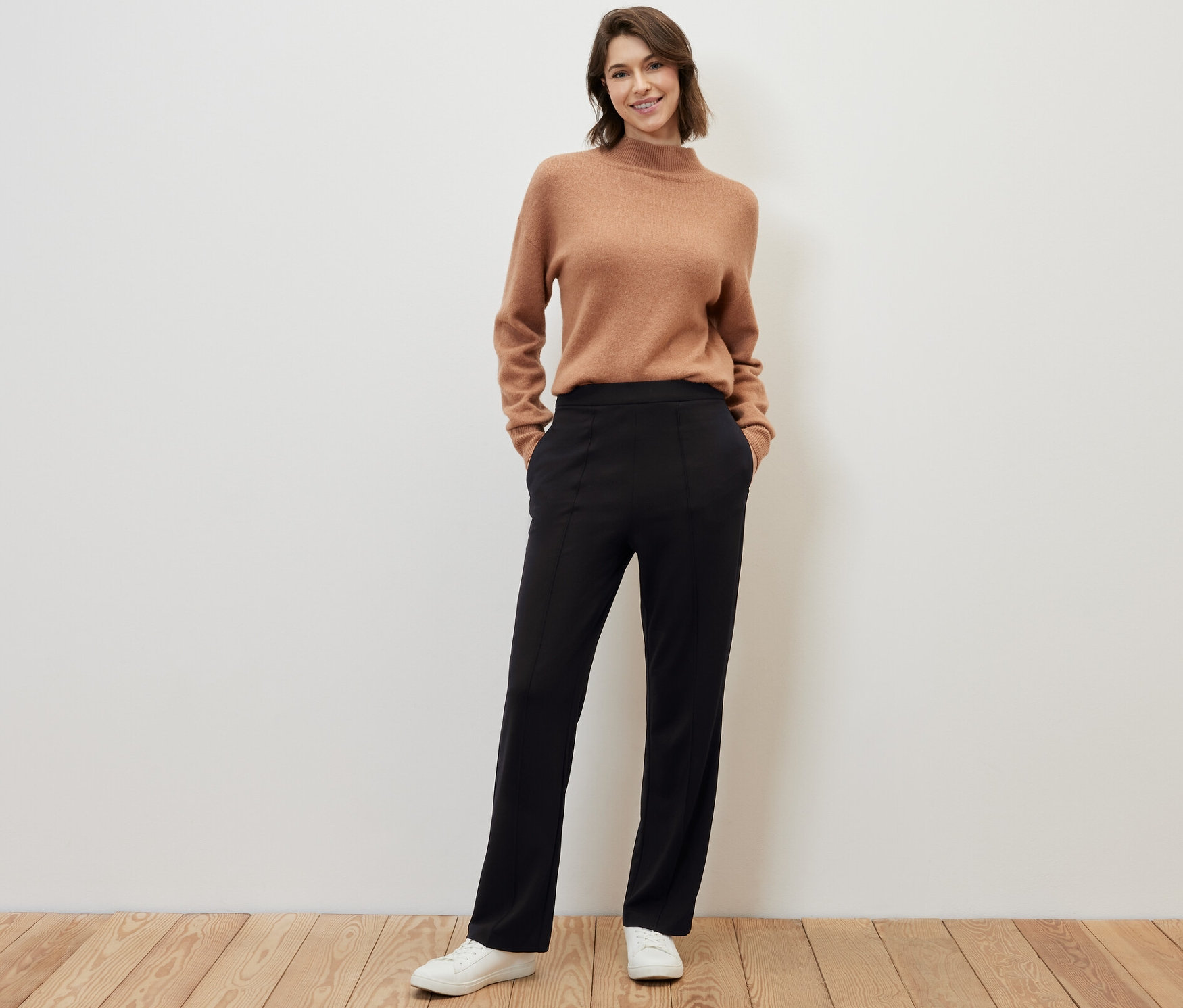 Femme posant sur un plancher en bois en pull marron et pantalon en jersey noir avec passepoil.