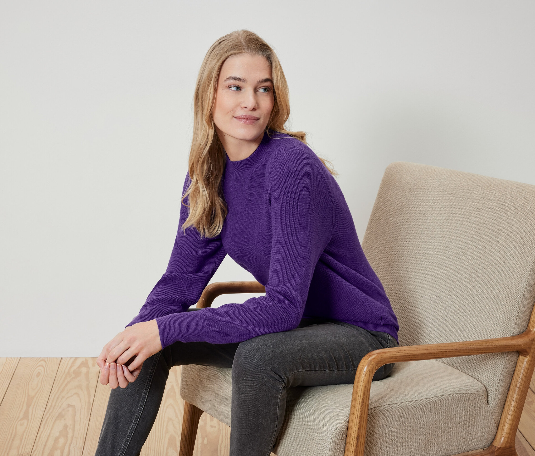 Une femme blonde est assise dans un fauteuil beige, porte un pull violet et un jean gris.