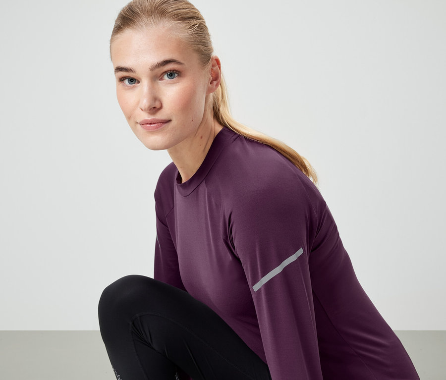 Une femme blonde portant un haut à manches longues violet et des leggings noirs regarde la caméra.