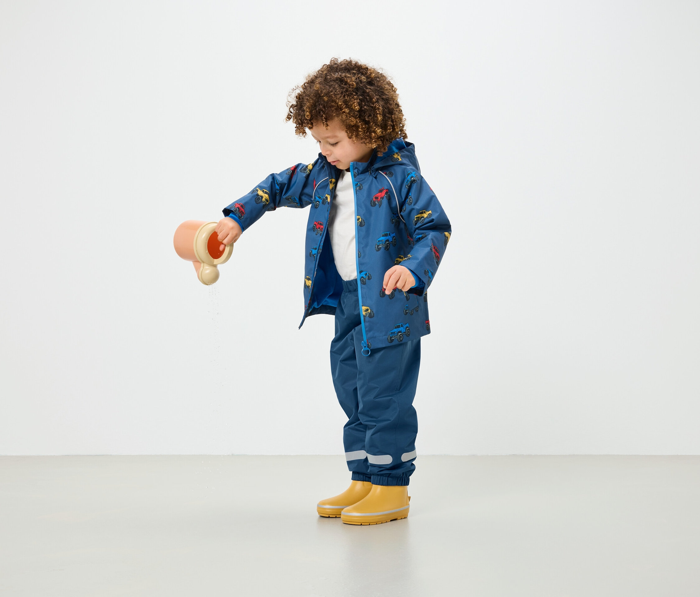 Un enfant aux cheveux bouclés verse de l'eau d'un jouet, portant un manteau de pluie bleu imprimé et un pantalon bleu.
