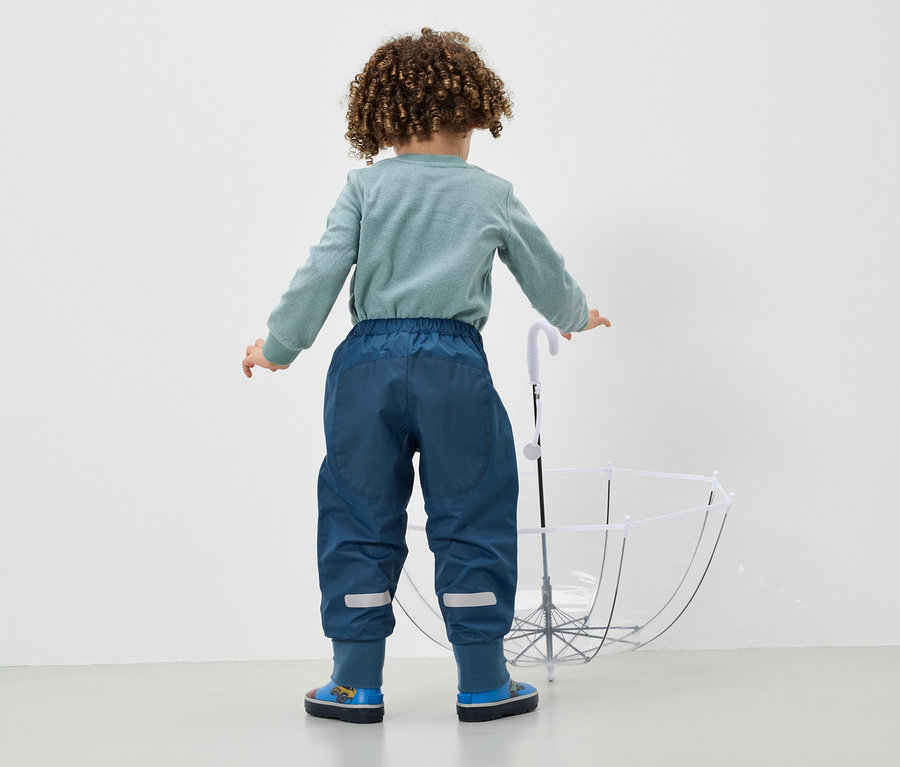 Un garçon en pantalon bleu et bottes bleues se tient dos à la caméra et tient un parapluie blanc.