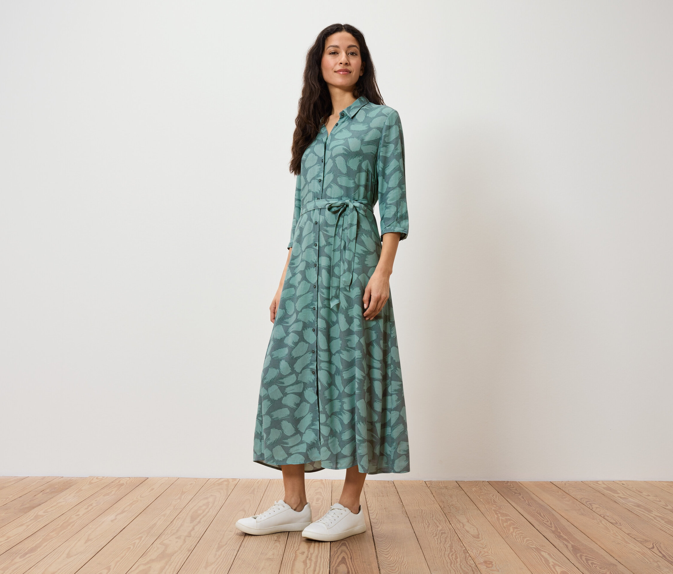 Une femme se tient debout et porte une robe chemise verte à imprimé et des baskets blanches.