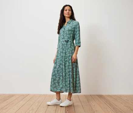 Une femme se tient debout et porte une robe chemise verte à imprimé et des baskets blanches.