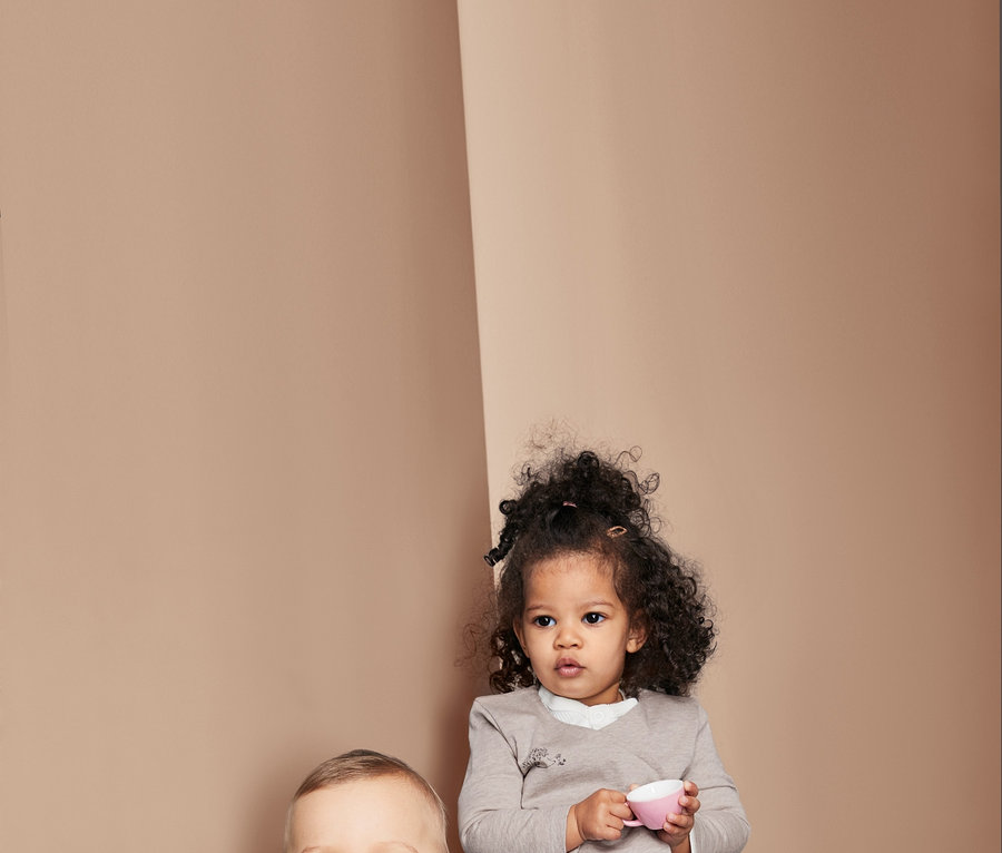 Deux enfants sont assis. Un garçon est assis sur un coussin et sourit, il porte un t-shirt rayé. Une fille tient une tasse rose et porte un haut. Tous deux portent des jeans.