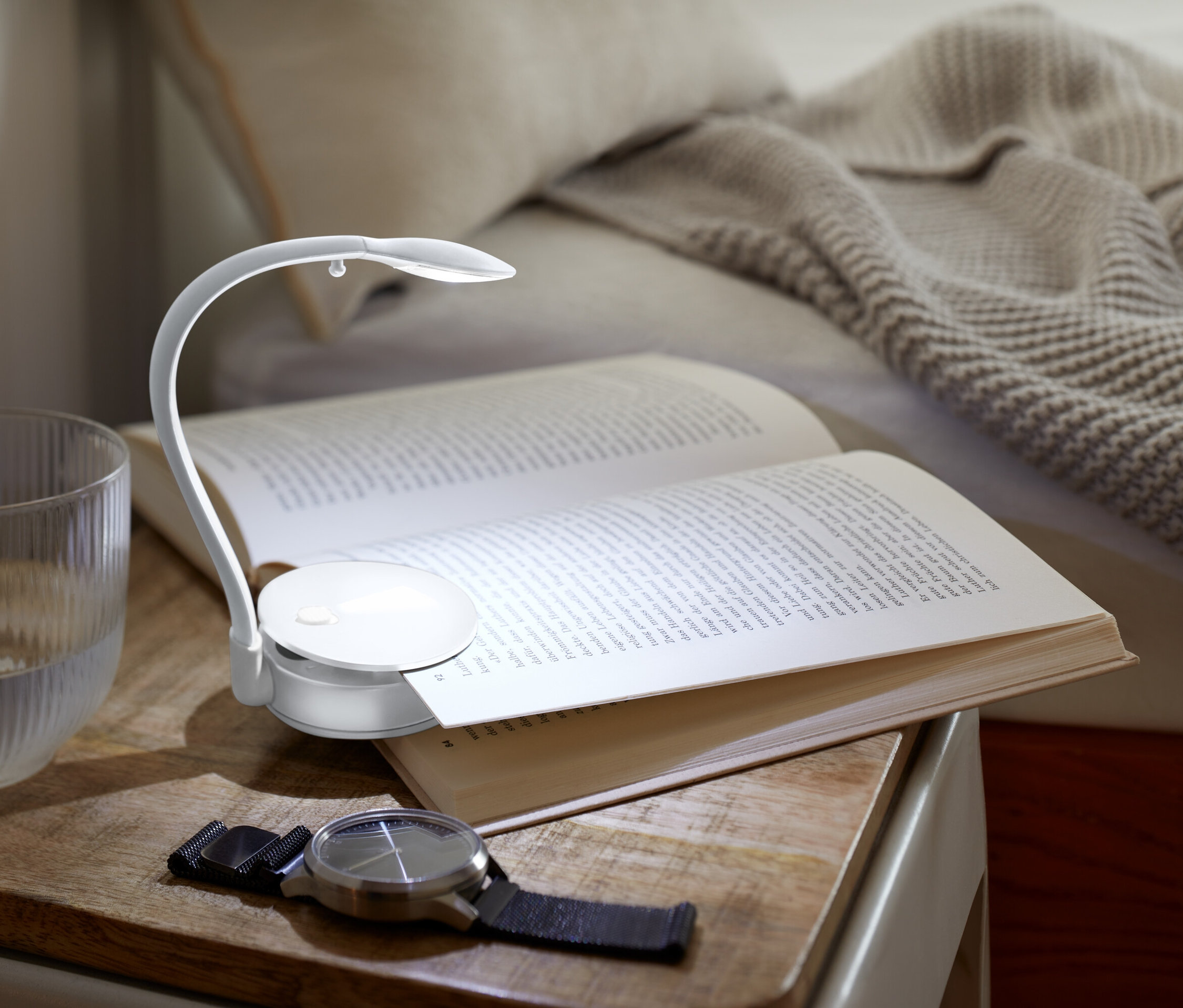 Sur une table de chevet se trouvent un livre ouvert éclairé par une petite lampe, un verre d'eau et une montre.