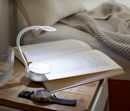 Sur une table de chevet se trouvent un livre ouvert éclairé par une petite lampe, un verre d'eau et une montre.