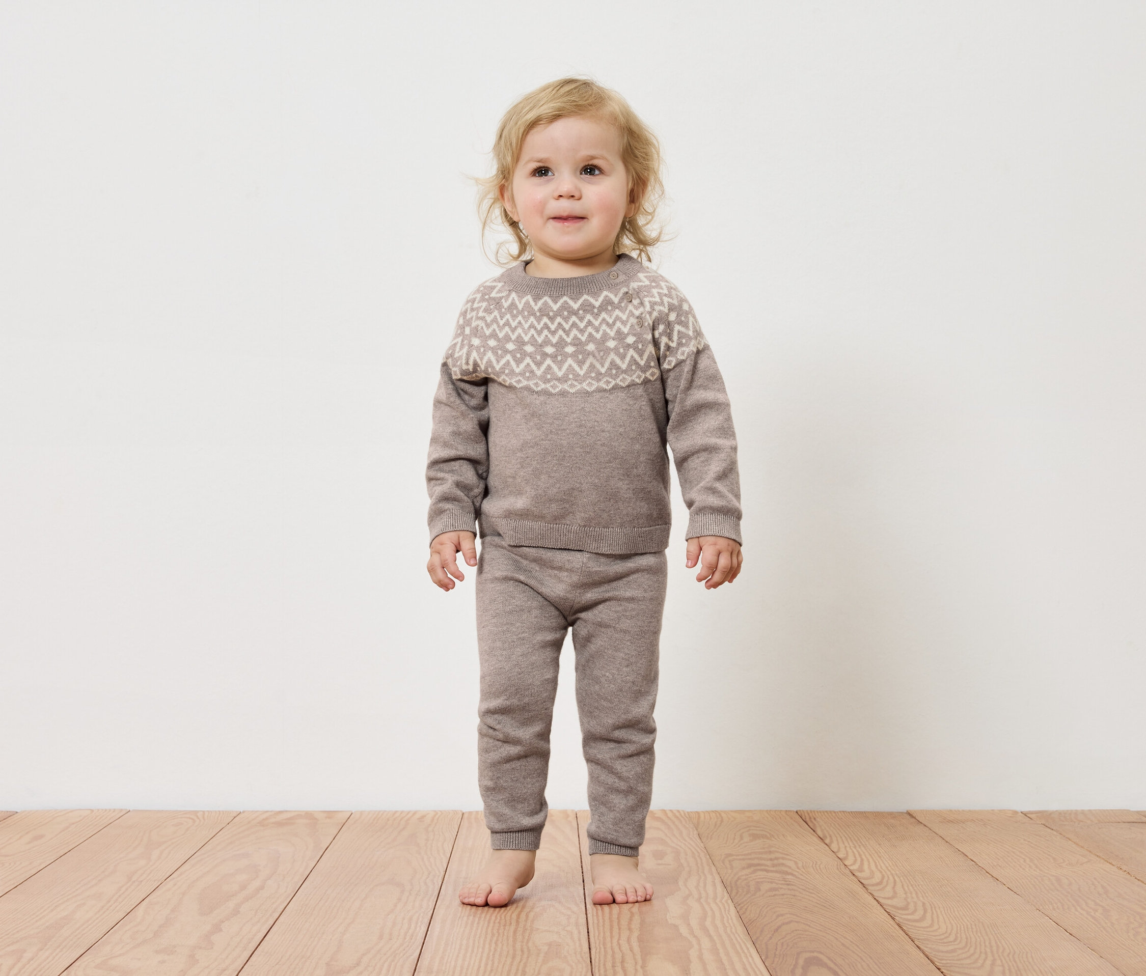 Un jeune enfant se tient sur un plancher de bois, portant un pull et un pantalon marron.