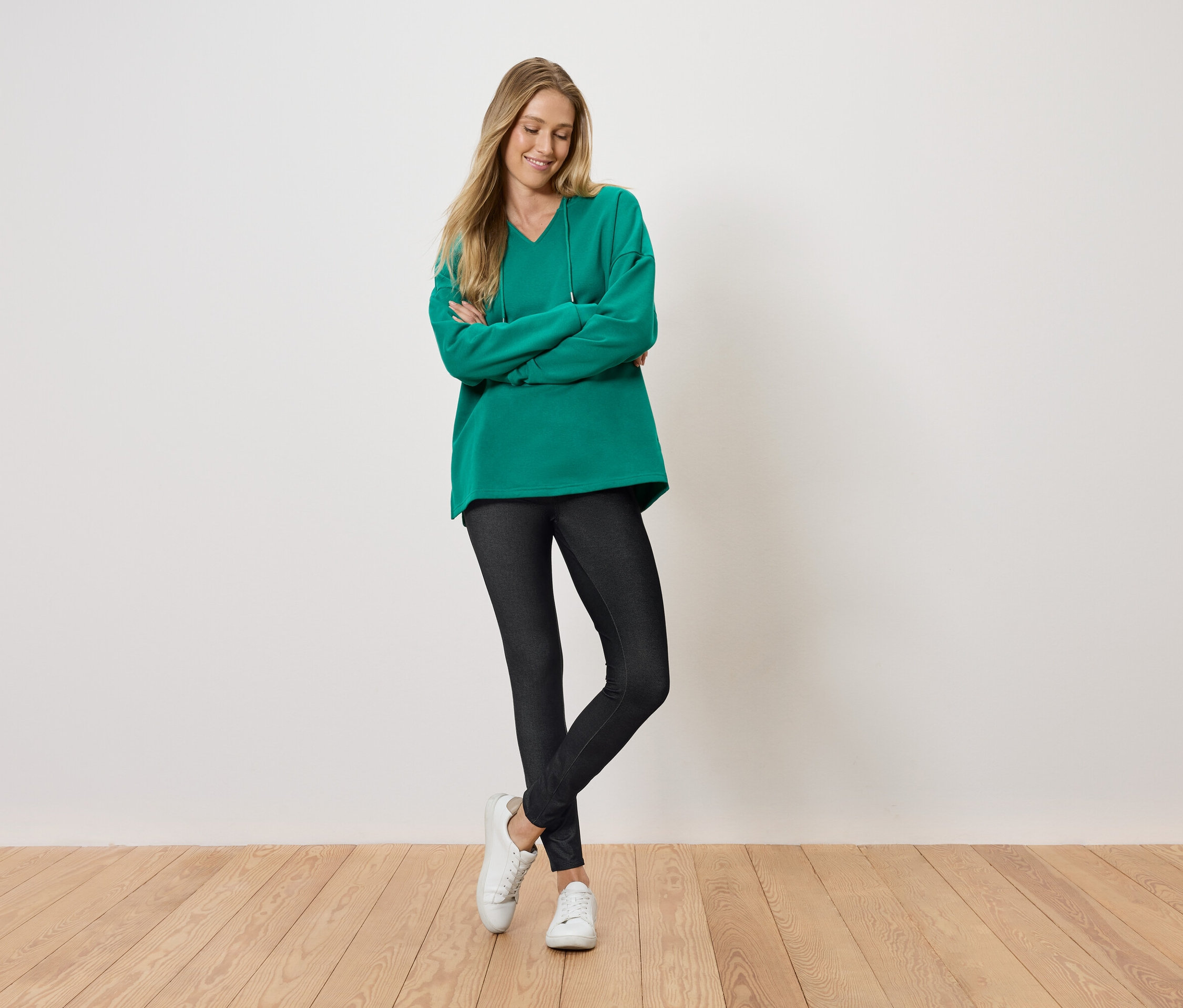 Une femme se tient debout, les bras croisés et souriant, portant un pull vert, un legging noir et des chaussures blanches.