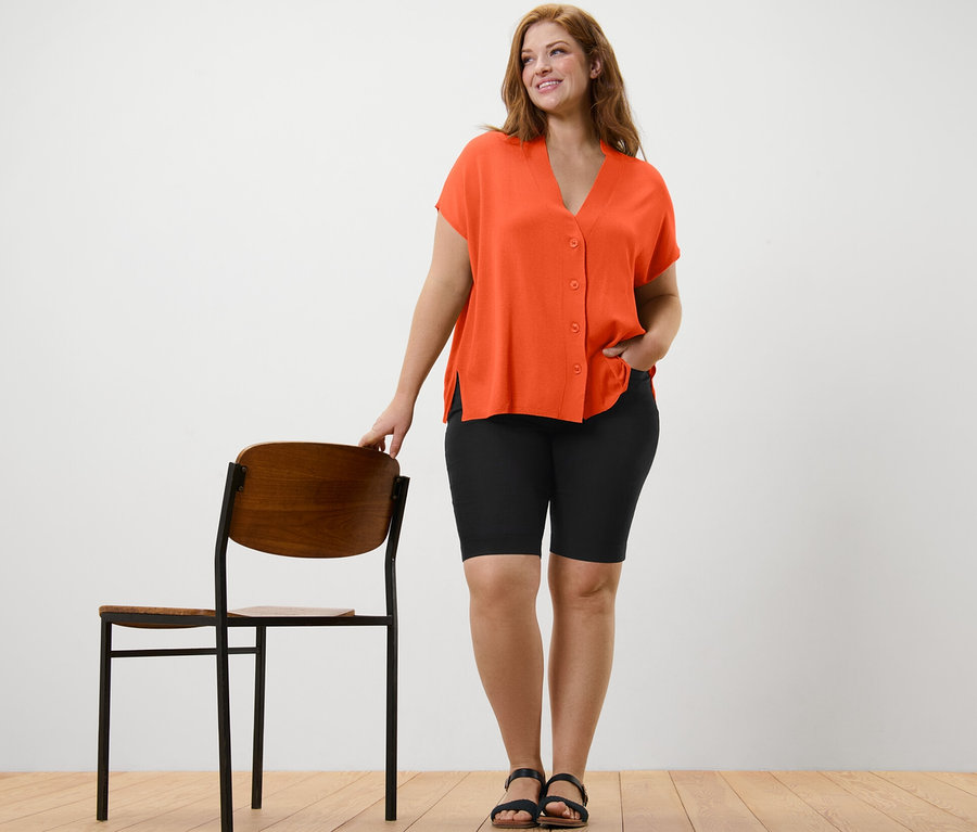 Une femme se tient à côté d'une chaise et porte un cardigan orange à manches courtes et un bermuda noir en bengaline.