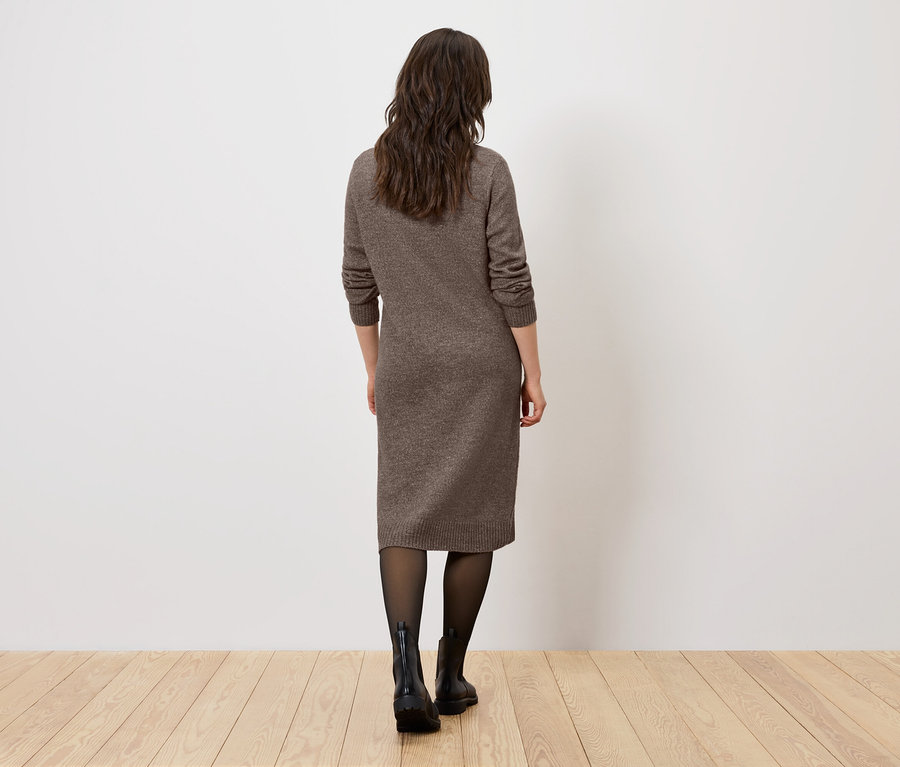 Une femme, dos à la caméra, se tient sur un plancher en bois et porte une robe marron, des collants noirs et des bottes noires.