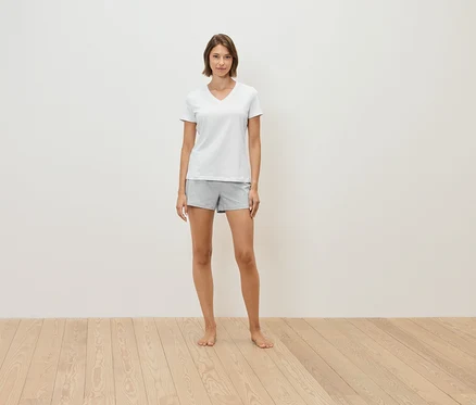 Une femme debout pieds nus sur un plancher en bois, portant un t-shirt blanc et un short gris.