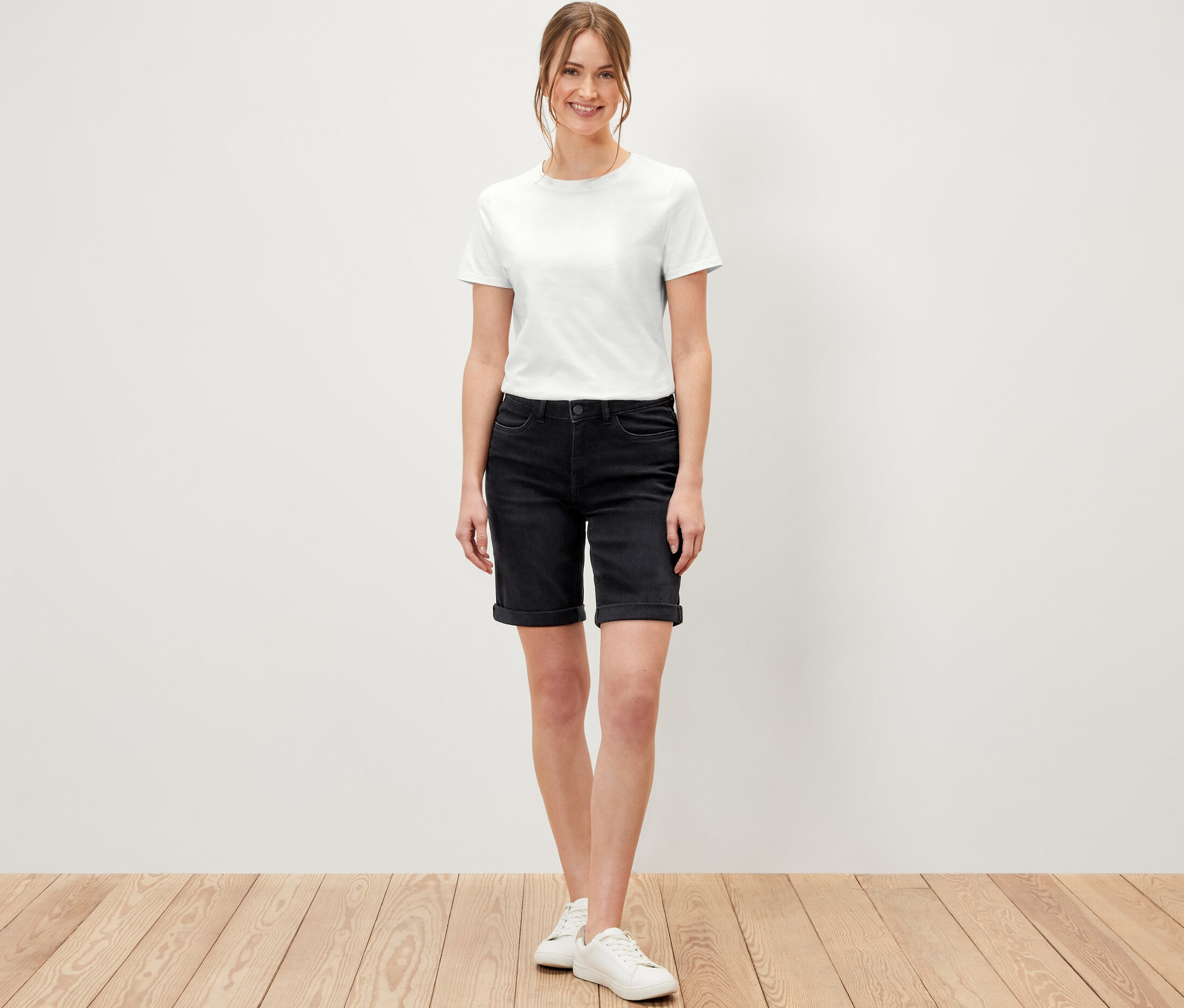 Une femme debout sur un plancher en bois porte un bermuda en jean coupe « Lea », denim noir et un t-shirt blanc.