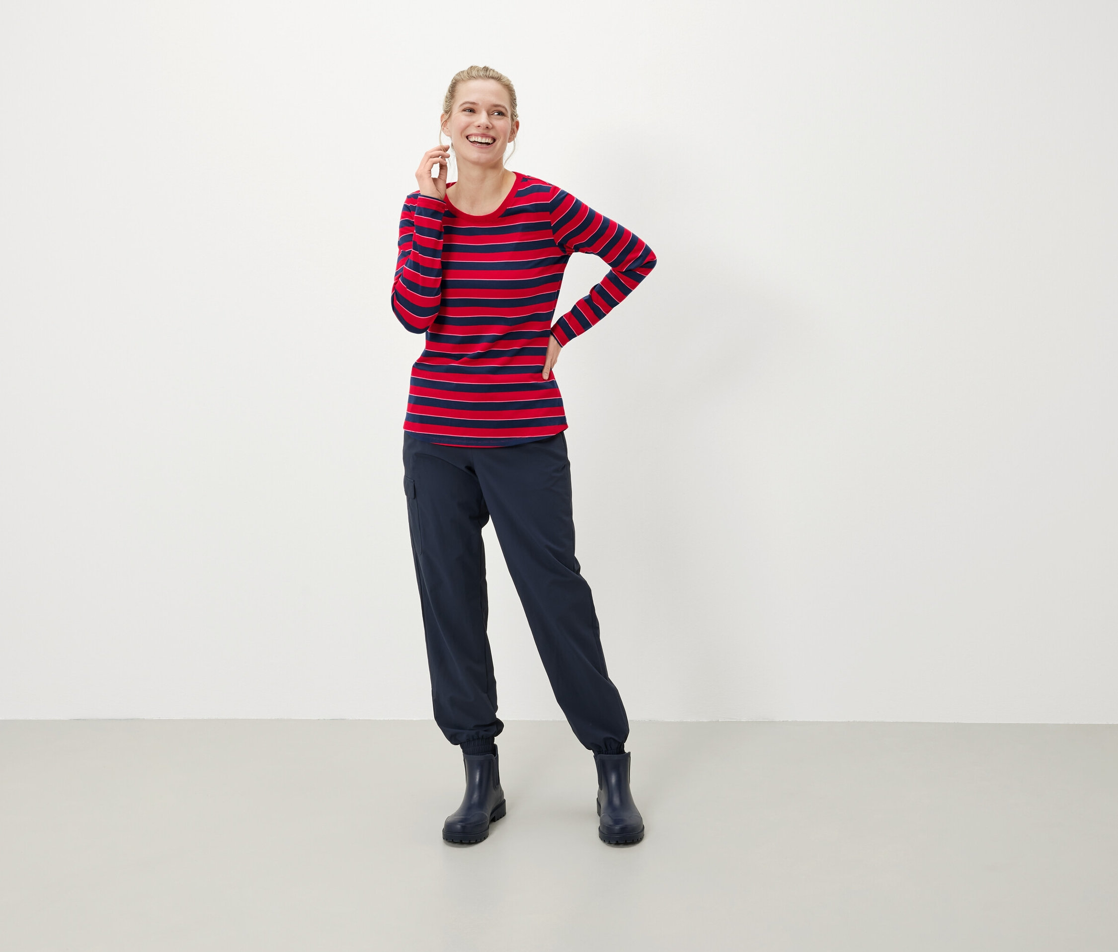 Une femme blonde sourit et tient un téléphone à son oreille. Elle porte un t-shirt à manches longues rayé rouge et bleu, un pantalon bleu foncé et des bottes.