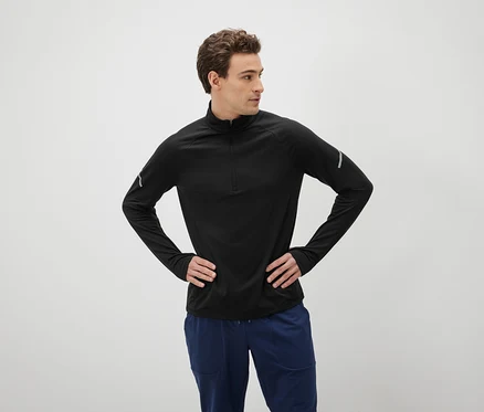 Un homme aux cheveux bruns pose les mains sur les hanches, vêtu d'un t-shirt noir à manches longues et d'un pantalon bleu.