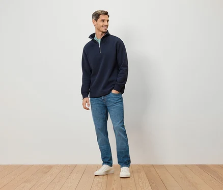 Un homme se tient devant un mur blanc. Il porte un pull bleu marine zippé, un jean bleu et des chaussures blanches.