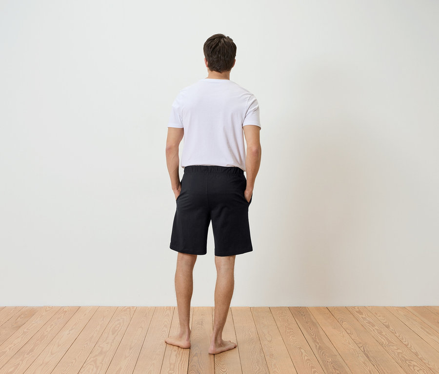 Un homme vu de dos, les mains dans les poches d'un short noir, portant un t-shirt blanc, se tenant sur un plancher en bois clair devant un mur blanc.