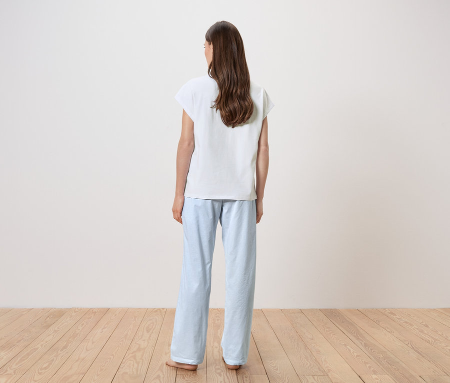 Femme debout, dos à la caméra, vêtue d'un t-shirt blanc et d'un pantalon de pyjama bleu clair.