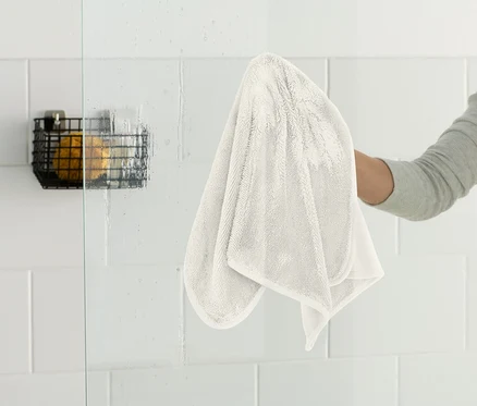 Une personne essuie une paroi de douche en verre avec un chiffon blanc. Un panier en fil de fer noir est accroché au mur.