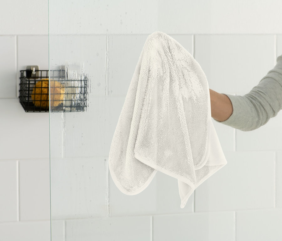 Une personne essuie une paroi de douche en verre avec un chiffon blanc. Un panier en fil de fer noir est accroché au mur.