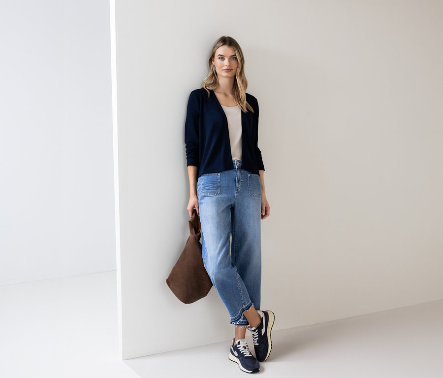 Une femme portant un cardigan bleu Street One, un jean et un sac marron est appuyée contre un mur.