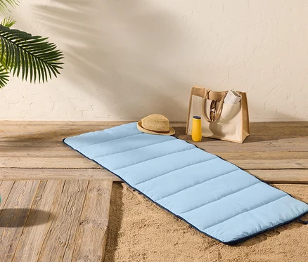 Un tapis de plage bleu clair est posé sur un plancher en bois à côté du sable, avec un chapeau de paille, un flacon de crème solaire et un sac fourre-tout à proximité.
