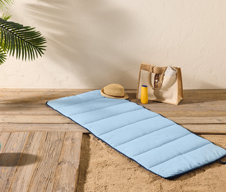 Un tapis de plage bleu clair est posé sur un plancher en bois à côté du sable, avec un chapeau de paille, un flacon de crème solaire et un sac fourre-tout à proximité.