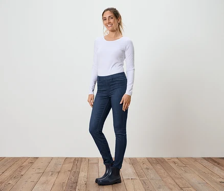 Une femme se tient de profil en jean, un T-shirt blanc à manches longues et des bottes en caoutchouc devant un mur blanc et un plancher en bois.
