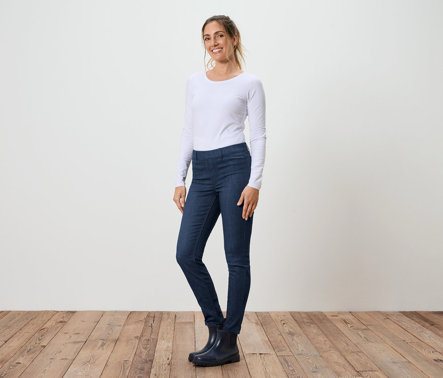 Une femme se tient de profil en jean, un T-shirt blanc à manches longues et des bottes en caoutchouc devant un mur blanc et un plancher en bois.