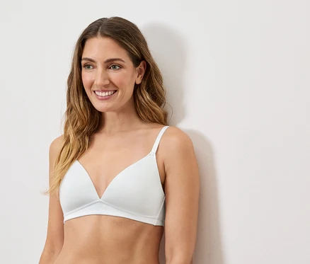 Une femme souriante aux cheveux ondulés porte un soutien-gorge triangle blanc à fines bretelles sur fond blanc.