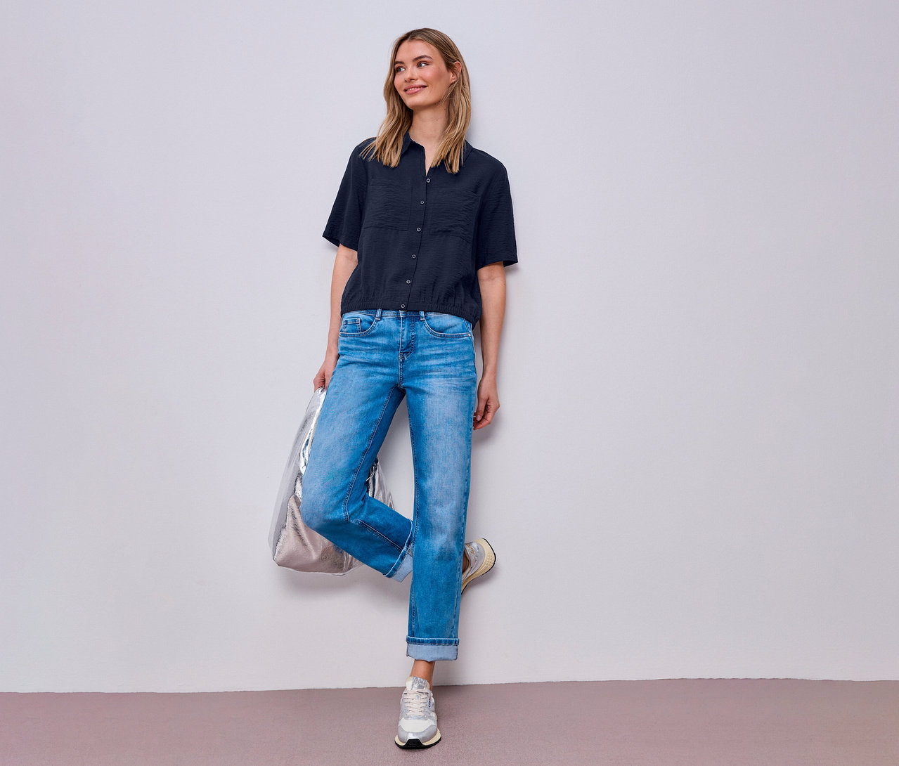 Une femme en jean et un chemisier Street One pose avec un sac argenté contre un mur.