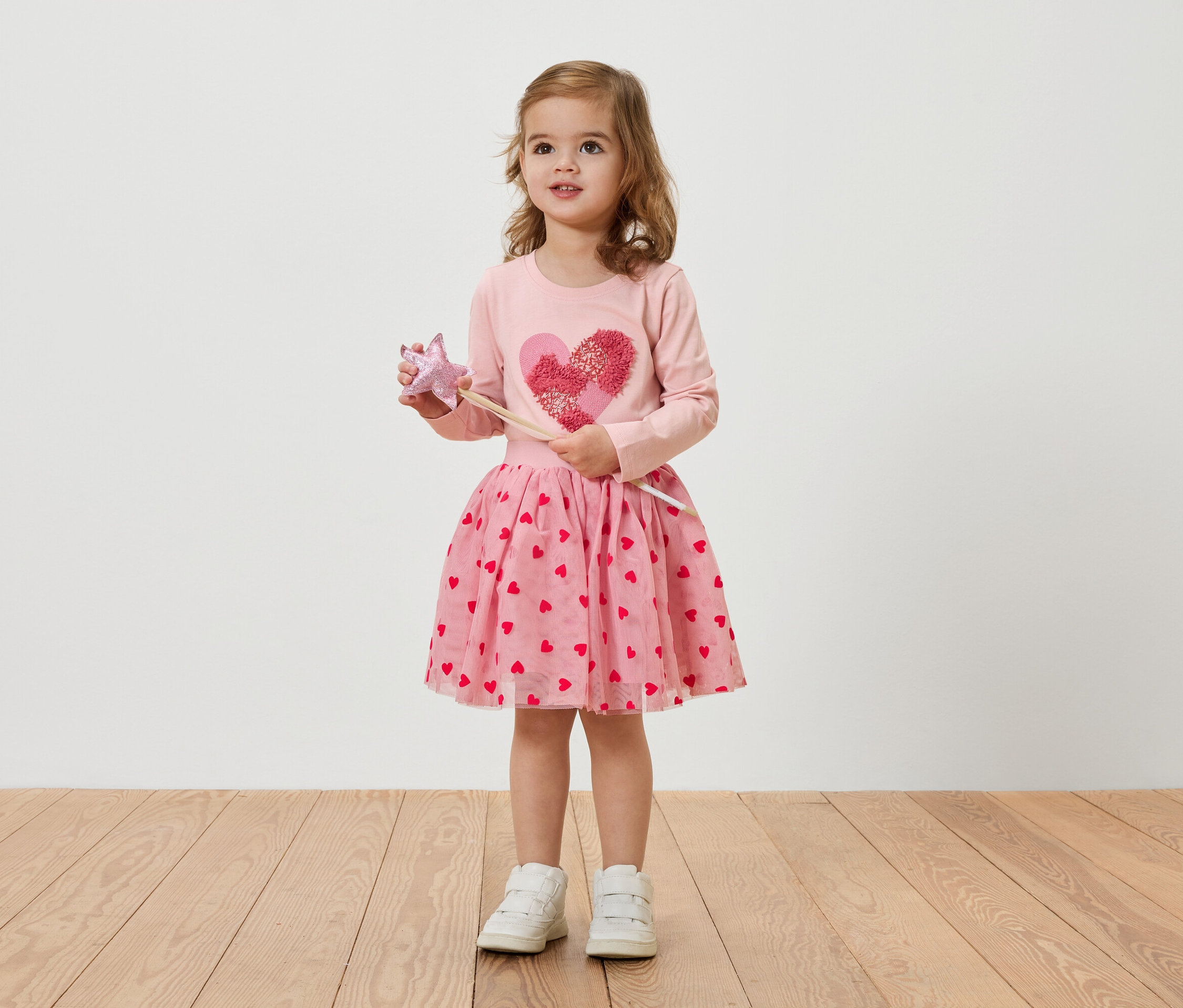 Une petite fille blonde porte une robe rose à motifs de coeurs et tient une baguette magique.