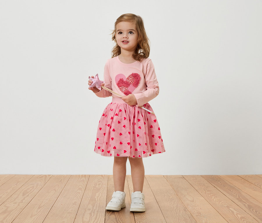 Une petite fille blonde porte une robe rose à motifs de coeurs et tient une baguette magique.