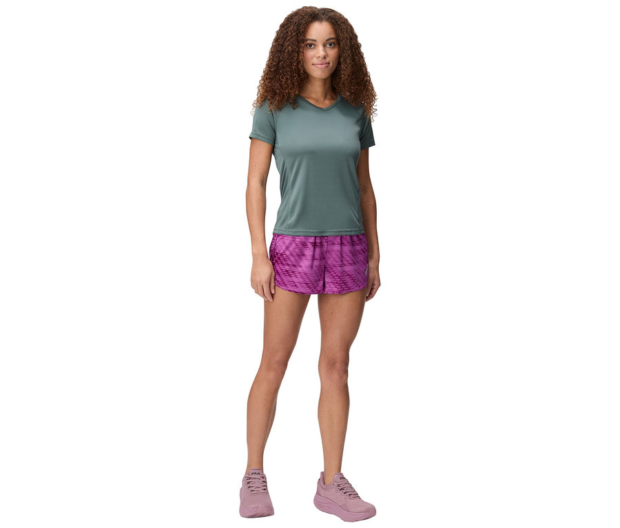 Femme debout sur fond blanc portant un t-shirt vert, un short rose et des chaussures roses.