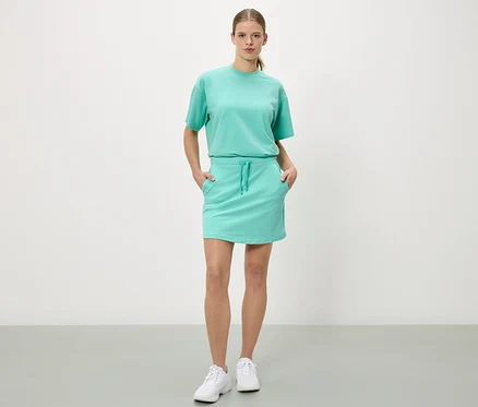 Femme debout, les mains dans les poches d'une jupe turquoise, avec un t-shirt assorti et des baskets blanches.