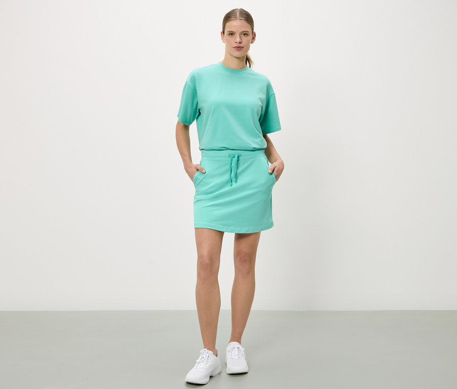 Femme debout, les mains dans les poches d'une jupe turquoise, avec un t-shirt assorti et des baskets blanches.