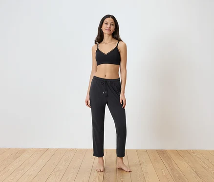 Une femme se tient pieds nus devant un mur blanc, vêtue d'un soutien-gorge noir en dentelle et d'un pantalon noir à cordon.