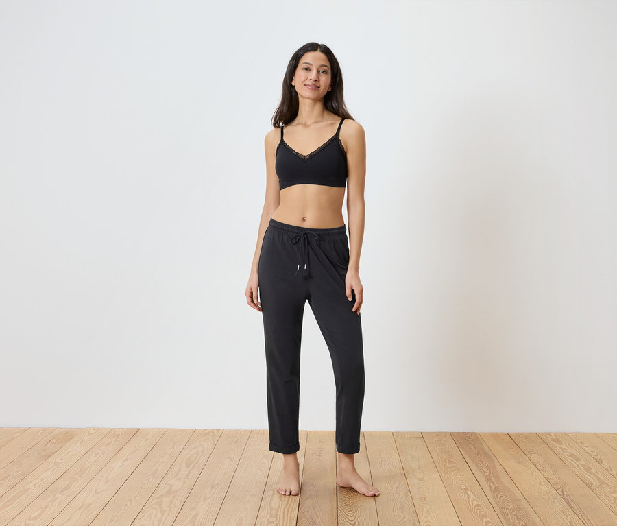 Une femme se tient pieds nus devant un mur blanc, vêtue d'un soutien-gorge noir en dentelle et d'un pantalon noir à cordon.